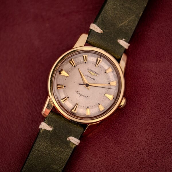 The Longines Automatic Conquest Ref. 9000-1 with a 14k gold capped case and green leather strap is showcased on a burgundy textured surface.