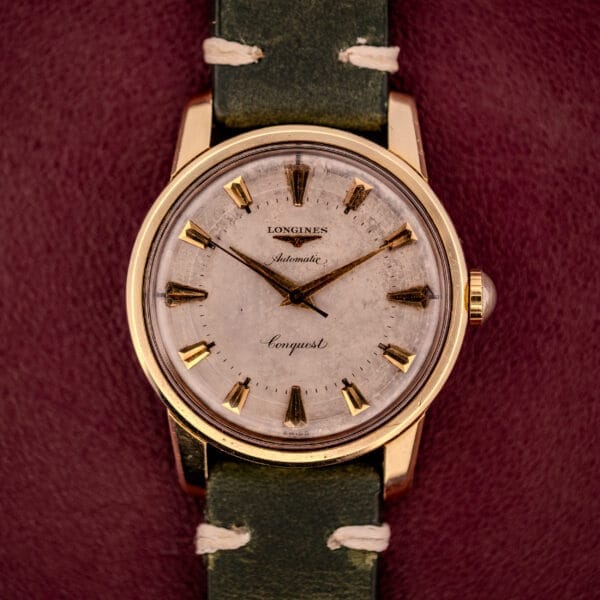 A Longines Automatic Conquest Ref. 9000-1 with a 14k gold capped case and green leather strap, pictured on a burgundy background.