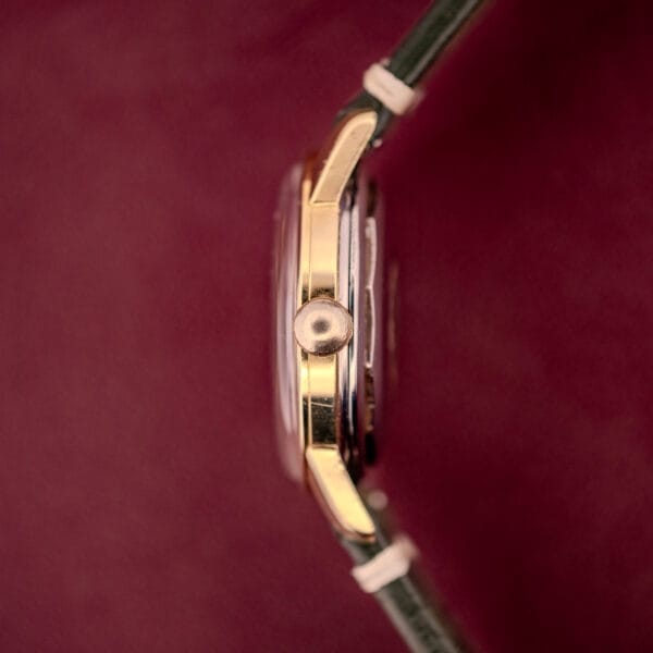 Side view of a Longines Automatic Conquest Ref. 9000-1 wristwatch with a 14k gold capped case and black strap, set against a burgundy background.