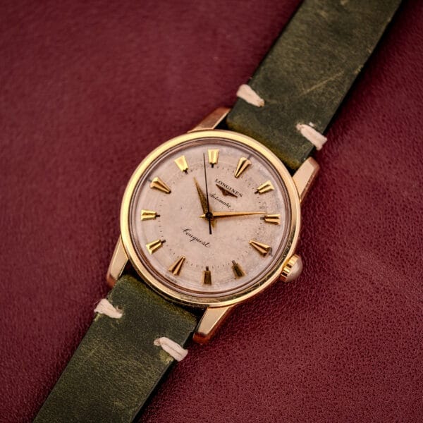 A Longines Automatic Conquest Ref. 9000-1 wristwatch featuring a 14k gold capped case, green leather strap, and textured dial set against a maroon background.