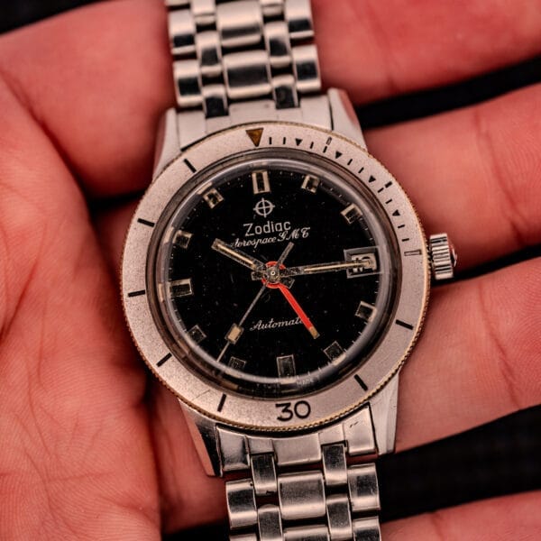 A hand holding a Zodiac Aerospace GMT Ref. 752-934 wristwatch with a chrome bezel, silver band, and black dial.