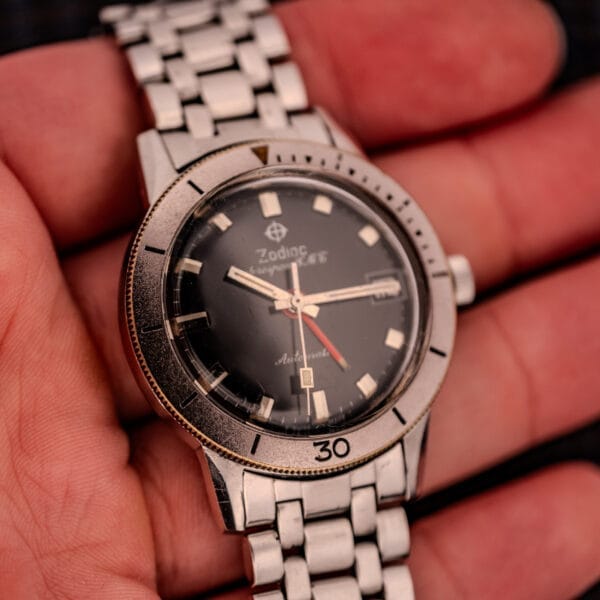A close-up of a hand holding a Zodiac Aerospace GMT Ref. 752-934 wristwatch featuring a black dial, chrome bezel, and metal band.
