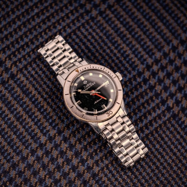 A Zodiac Aerospace Gmt Ref. 752-934 Black Dial & Chrome Bezel wristwatch with a metal band lies on blue and brown houndstooth fabric.