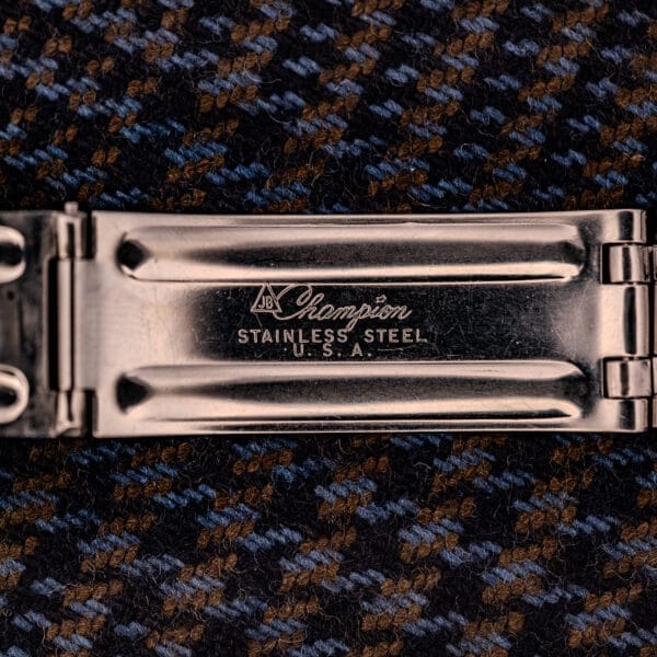 Close-up of a metal watch clasp engraved with Champion Stainless Steel U.S.A., set against patterned fabric, beside the Zodiac Aerospace GMT Ref. 752-934 with black dial and chrome bezel.