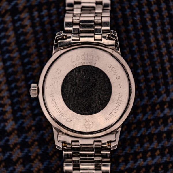 A close-up of the back of a Zodiac Aerospace GMT Ref. 752-934 with black dial and chrome bezel, featuring engraved text, resting on patterned fabric.