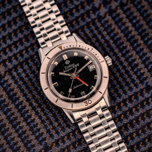 A Zodiac Aerospace GMT Ref. 752-934 wristwatch with a black dial and chrome bezel is displayed on blue and brown patterned fabric.