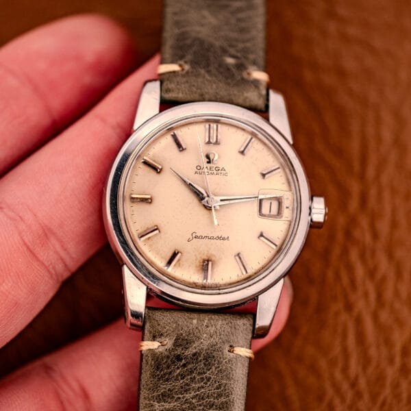 A hand holds an Omega Automatic Seamaster Ref. 2849 Stainless Steel Chunky Lugs featuring a tan dial and green leather strap.