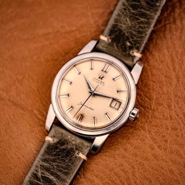 An Omega Automatic Seamaster Ref. 2849 Stainless Steel Chunky Lugs watch with a tan dial and dark leather strap rests elegantly on a brown leather surface.