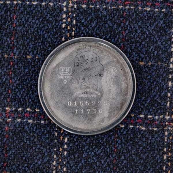 A vintage metal case back with engraved numbers and handwritten notes, likely from a Movado "Acvatic" FB Case Sector Dial Ref. 11730 Cal. 150MN, rests on blue plaid fabric.