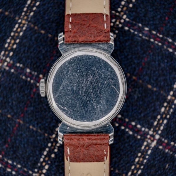 A Movado "Acvatic" FB Case Sector Dial Ref. 11730 Cal. 150MN watch with a scratched back and brown leather strap rests on blue plaid fabric.