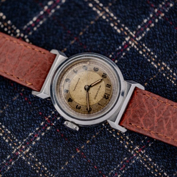 A vintage Movado "Acvatic" FB Case Sector Dial Ref. 11730 Cal. 150MN with a brown leather strap rests elegantly on a dark plaid fabric background.
