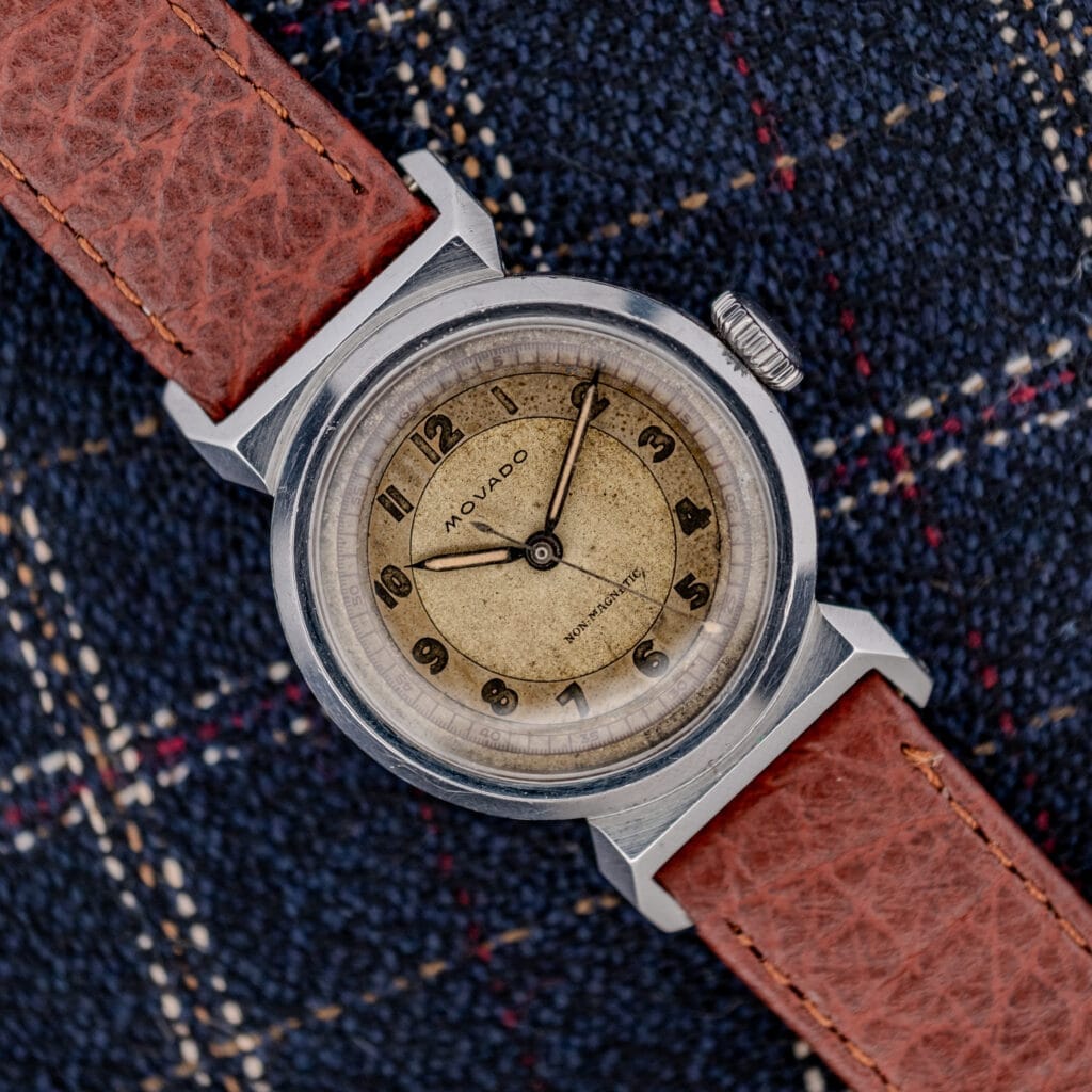 A Movado "Acvatic" FB Case Sector Dial Ref. 11730 Cal. 150MN wristwatch with a brown leather strap rests on a blue plaid fabric background.