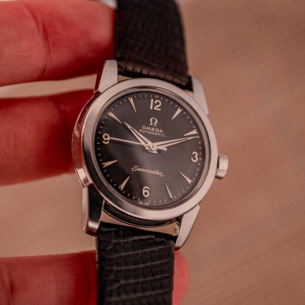 A hand holding a 1956 Omega Seamaster Automatic Ref. 2828-6 SC Cal. 470 Stainless Steel wristwatch with a black leather strap.