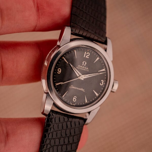 A hand holds a 1956 Omega Seamaster Automatic Ref. 2828-6 SC Cal. 470 wristwatch in stainless steel with a black dial and textured black leather strap.