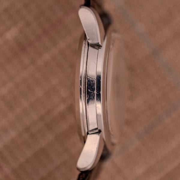 Side view of a 1956 Omega Seamaster Automatic Ref. 2828-6 SC Cal. 470 Stainless Steel case with visible scratches, set against a brown textured background.