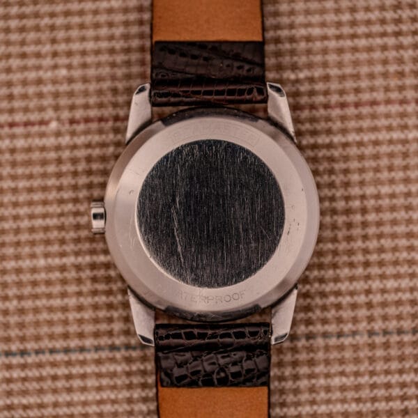 The back of a 1956 Omega Seamaster Automatic Ref. 2828-6 SC Cal. 470 stainless steel wristwatch with a brown leather strap rests on a checkered fabric background.