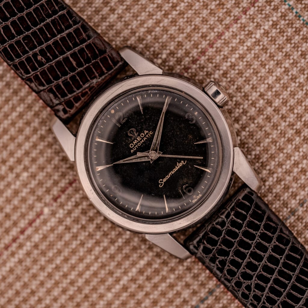 1956 Omega Seamaster Automatic Ref. 2828-6 SC Cal. 470 Stainless Steel with black dial and brown textured leather strap, shown on a patterned fabric background.