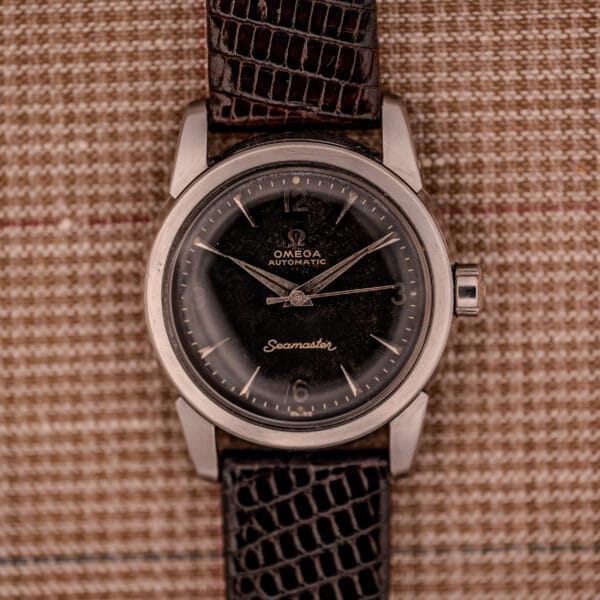A 1956 Omega Seamaster Automatic Ref. 2828-6 SC Cal. 470 in stainless steel with a black dial and brown leather strap, displayed on plaid fabric.