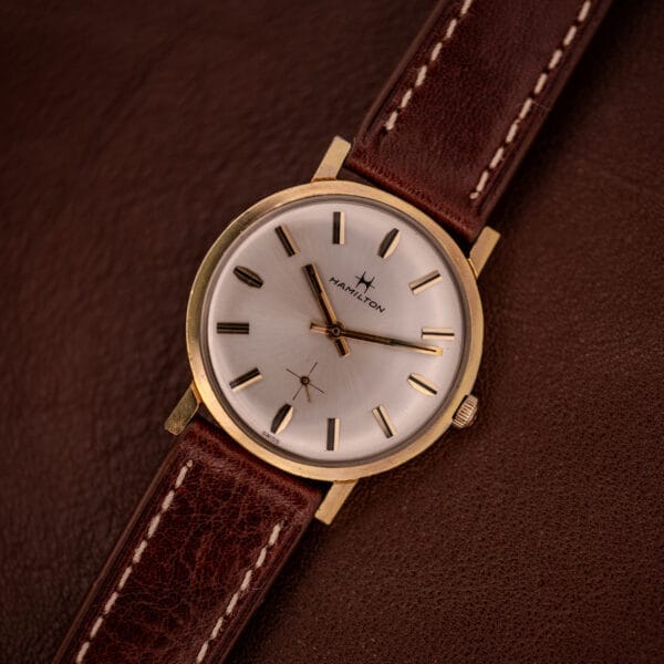 The Hamilton Manual Wind Cal. 638, featuring a gold tone case, sunburst dial, and brown leather strap, is displayed elegantly on a matching leather surface.