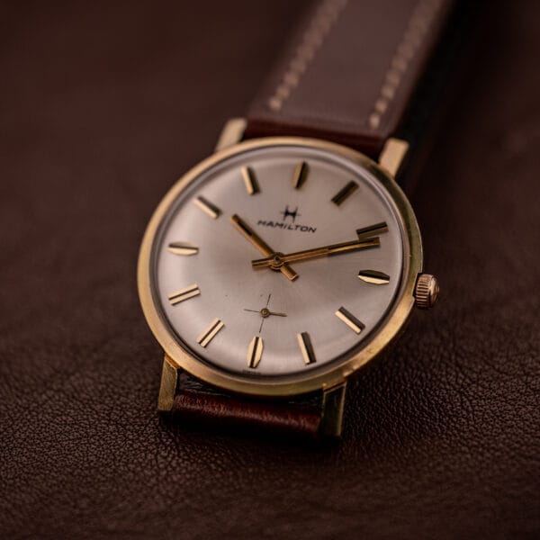 The Hamilton Manual Wind Cal. 638 Sunburst Dial Gold Tone Case wristwatch, with a brown leather strap, is elegantly displayed on a matching brown leather surface.