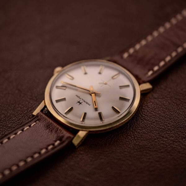 A Hamilton Manual Wind Cal. 638 Sunburst Dial gold tone case wristwatch with a brown leather strap rests on a brown textured surface.
