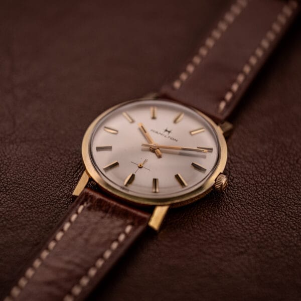 The Hamilton Manual Wind Cal. 638 features a sunburst dial and gold tone case, paired with a brown leather strap and elegantly displayed on a matching brown leather surface.