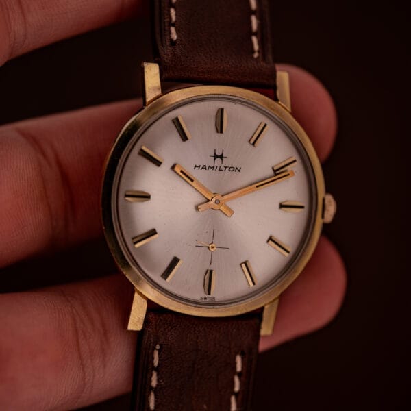 A hand holds the Hamilton Manual Wind Cal. 638 Sunburst Dial Gold Tone Case wristwatch with a brown leather strap, displaying the time 1:40.