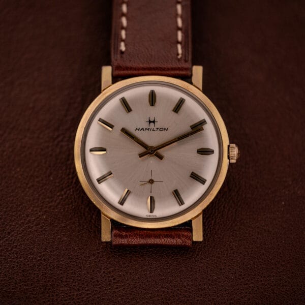 The Hamilton Manual Wind Cal. 638 Sunburst Dial Gold Tone Case wristwatch with a brown leather strap rests on a textured brown surface, highlighting its elegant vintage design.