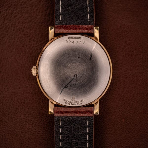 The back of a Hamilton Manual Wind Cal. 638 Sunburst Dial wristwatch with a gold tone case and brown leather strap rests on a brown surface.
