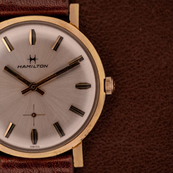 Close-up of a Hamilton Manual Wind Cal. 638 wristwatch with sunburst dial, gold tone case, and brown leather strap, set against a brown background.