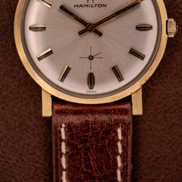 Close-up of the Hamilton Manual Wind Cal. 638 with a sunburst dial, gold tone case, and brown leather strap, displayed on a brown background.