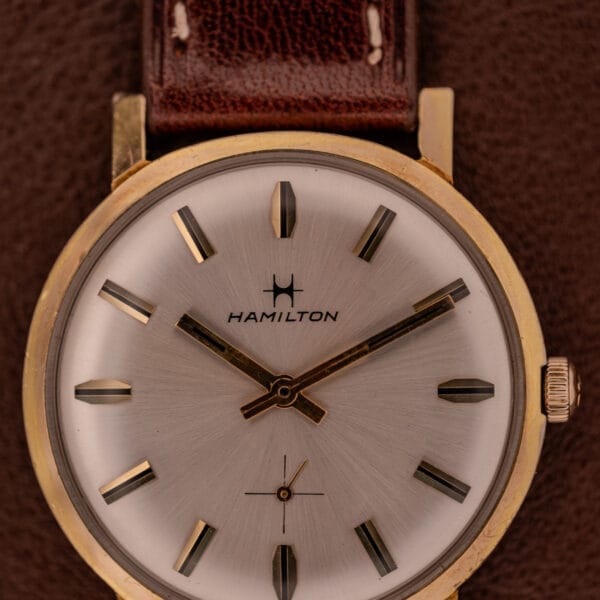 Close-up of a Hamilton Manual Wind Cal. 638 wristwatch featuring a gold tone case, sunburst dial, and brown leather band on a brown textured background.