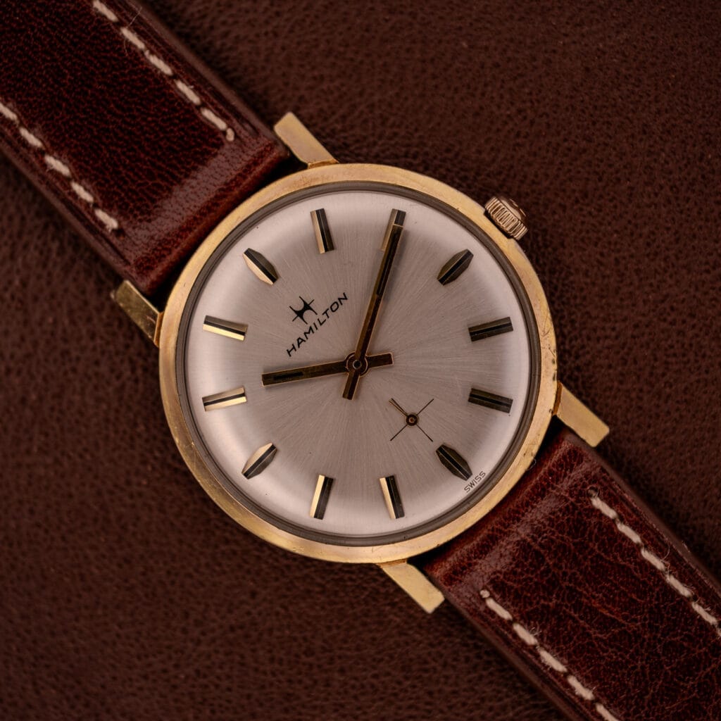 The Hamilton Manual Wind Cal. 638 Sunburst Dial Gold Tone Case watch with a brown leather strap rests on a brown textured surface.