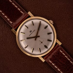 The Hamilton Manual Wind Cal. 638 Sunburst Dial Gold Tone Case watch with a brown leather strap rests on a brown textured surface.