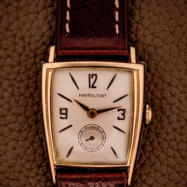 A Hamilton Asymmetric Khyber Manual Wind Caliber 730 wristwatch with a brown leather strap is displayed on a textured brown background.