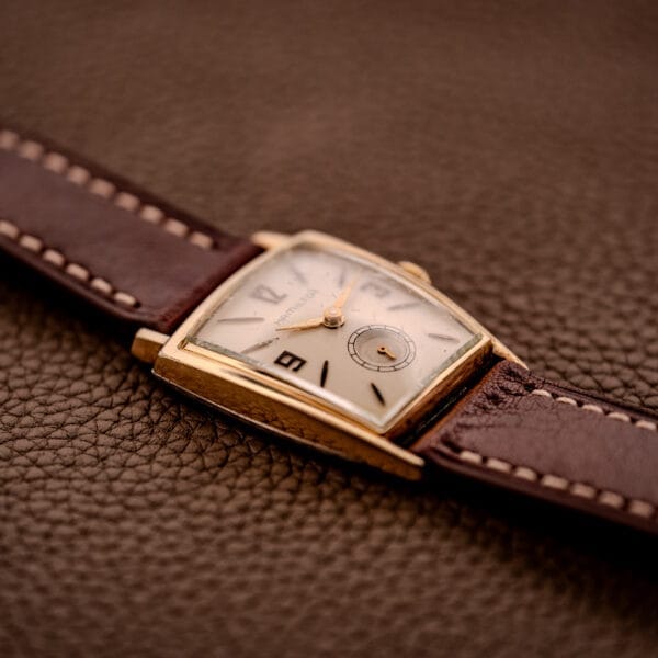 Hamilton Asymmetric Khyber Manual Wind Caliber 730 wristwatch with an angled gold case and brown leather strap, displayed on a textured brown surface.