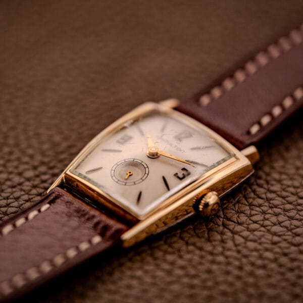 A vintage Hamilton Asymmetric Khyber Manual Wind Caliber 730 wristwatch with a brown leather strap is displayed on a textured brown surface.