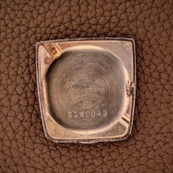 Close-up of the Hamilton Asymmetric Khyber Manual Wind Caliber 730 metal case back, featuring engraved text and caliber number, resting on textured brown leather.