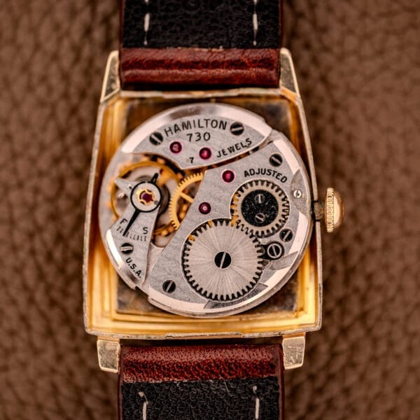 Close-up of the Hamilton Asymmetric Khyber Manual Wind Caliber 730 watch movement with visible gears and jewels, set against a textured brown background.
