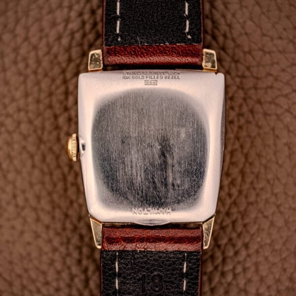 The back of a Hamilton Asymmetric Khyber Manual Wind Caliber 730 wristwatch, with a worn silver case and black leather strap, rests on a textured brown surface.
