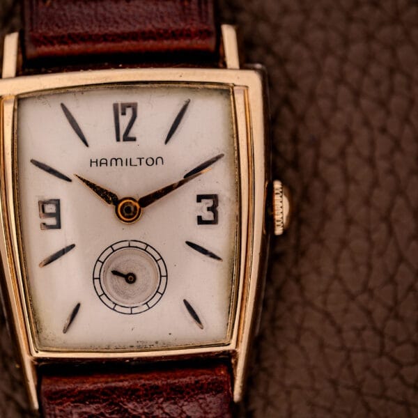 Close-up of a vintage Hamilton Asymmetric Khyber Manual Wind Caliber 730 wristwatch with a square face and brown leather strap on a textured surface.