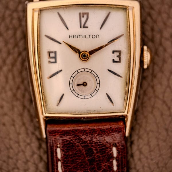 A Hamilton Asymmetric Khyber wristwatch with a rectangular face, brown leather strap, and Manual Wind Caliber 730 movement is displayed on a textured surface.