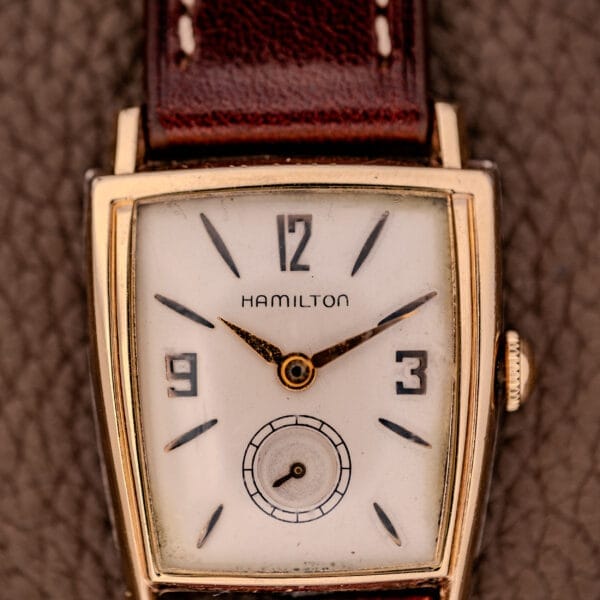 The Hamilton Asymmetric Khyber Manual Wind Caliber 730 wristwatch features a gold rectangular case and brown leather strap, shown against a textured brown background.