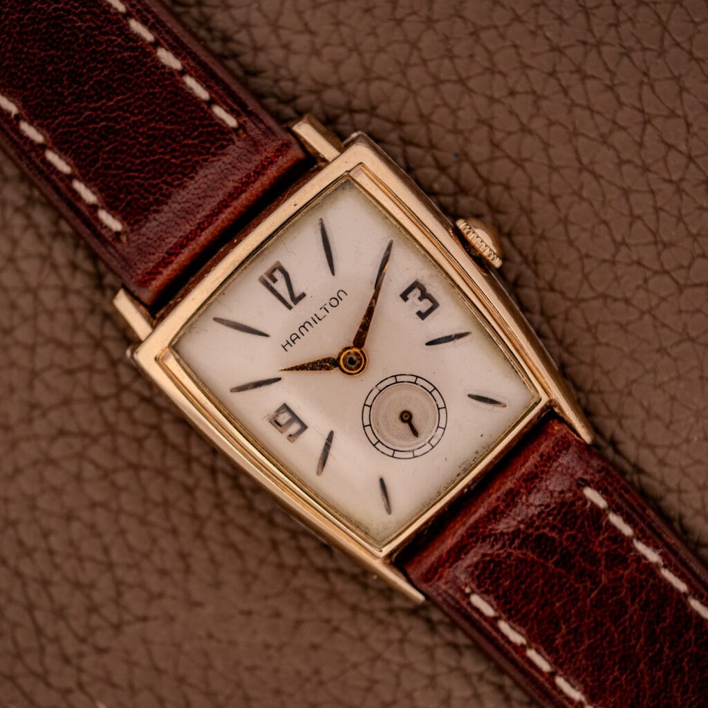 The Hamilton Asymmetric Khyber Manual Wind Caliber 730 wristwatch, with a rectangular face and brown leather strap, rests elegantly on a textured brown surface.