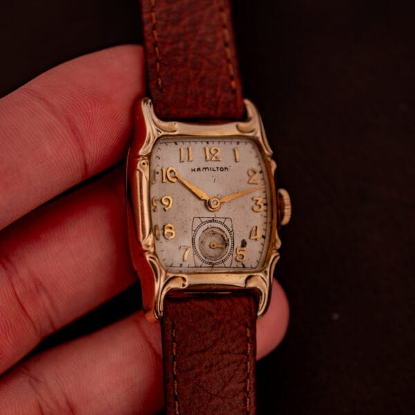 A hand holds a Hamilton Lawton Art Deco Gold Filled Case Manual Wind Cal. 747 wristwatch with a square face and brown leather strap.