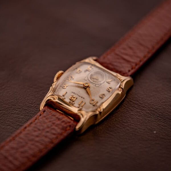 The Hamilton Lawton Art Deco Gold Filled Case Manual Wind Cal. 747 wristwatch, featuring a luxurious gold filled case and brown leather strap, rests on a dark surface, showcasing its timeless vintage elegance.