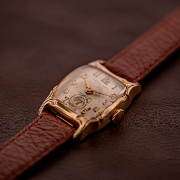 A Hamilton Lawton Art Deco Gold Filled Case Manual Wind Cal. 747 wristwatch with a brown leather strap rests elegantly on a brown surface.