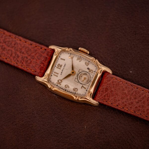 The Hamilton Lawton Art Deco Gold Filled Case Manual Wind Cal. 747 wristwatch features a gold filled case and brown leather strap, displayed on a dark brown leather surface.