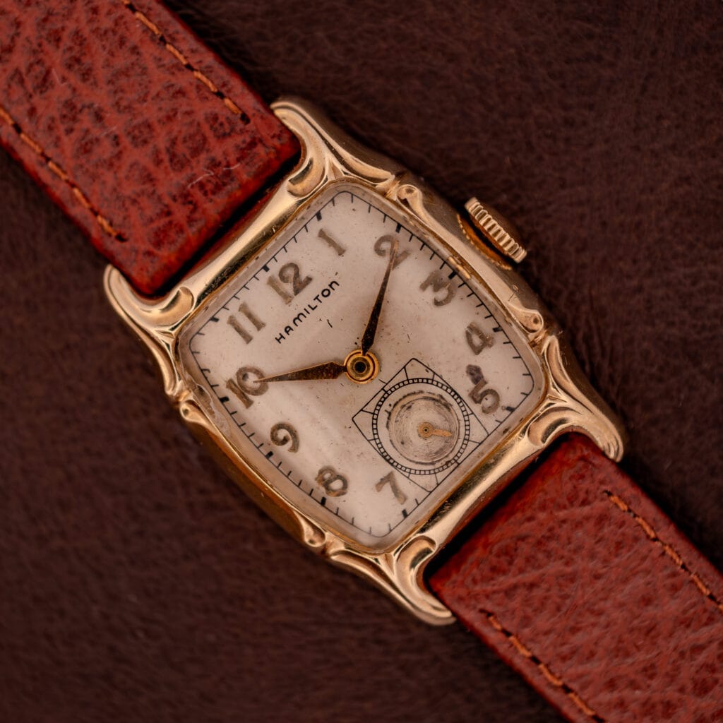 Hamilton Lawton Art Deco wristwatch with a gold filled case, manual wind Cal. 747 movement, white dial, and brown leather strap displayed on a brown surface.