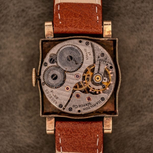 Close-up of a Hamilton Art Deco Square Gold Filled Case Manual Wind Cal. 987-F watch with visible gears and a brown leather strap on a textured surface.
